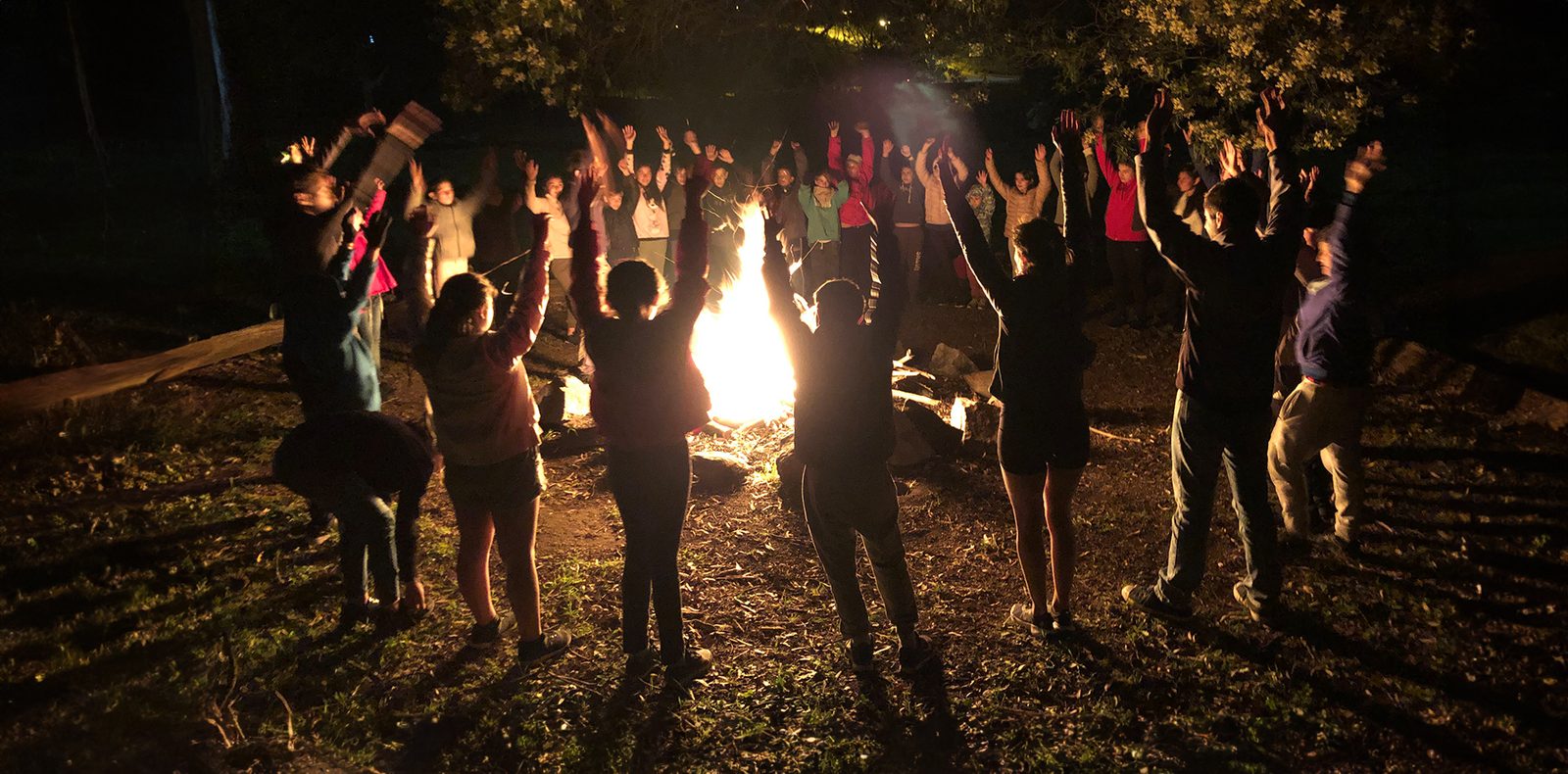 Grupo de chicos en fogón nocturno con manos arriba en Tandil Aventura